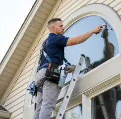 Professional window cleaner on a ladder using a squeegee to clean an exterior arched window, Arvada, CO.